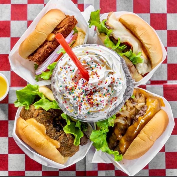 Four Halalbee's burgers and a milkshake with whipped cream and sprinkles on a checkered tablecloth.