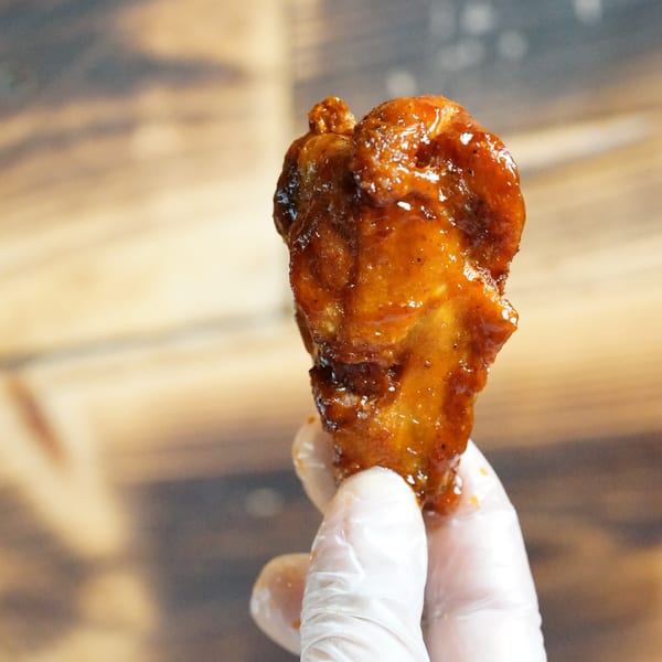A gloved hand holds up a single glistening Halalbee's chicken wing against a wooden background.