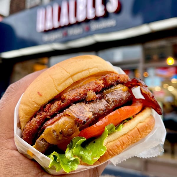 A hand holds a Halalbee's burger with a crispy patty, cheese, lettuce, and tomato, in front of the restaurant.