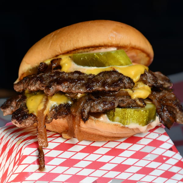 A Halalbee's double smash burger with melted cheese, caramelized onions, and pickles on a red and white checkered paper.